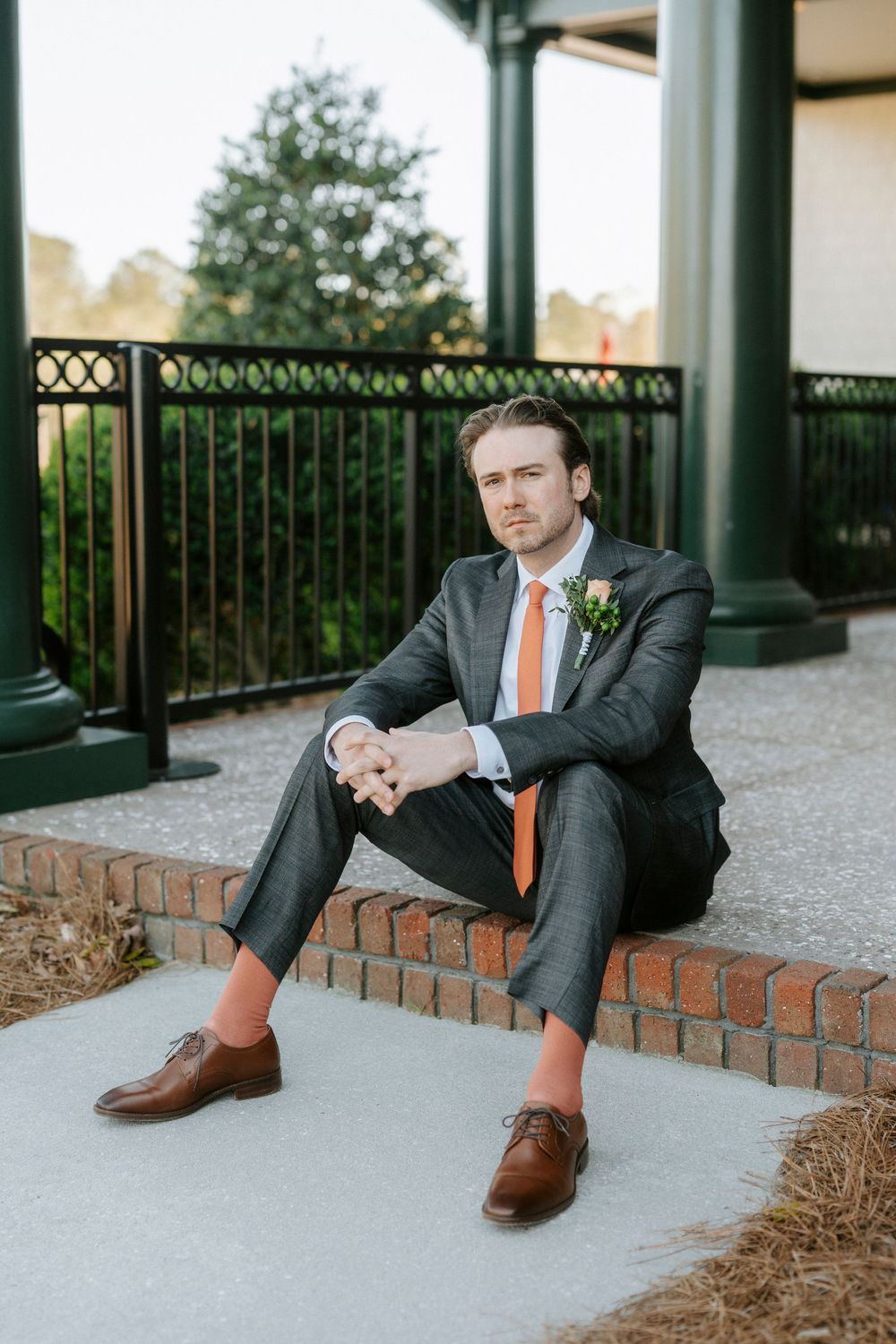 Close-up details of brown leather dress shoes and gray suit pants while sitting on brick ledge at outdoor venue.