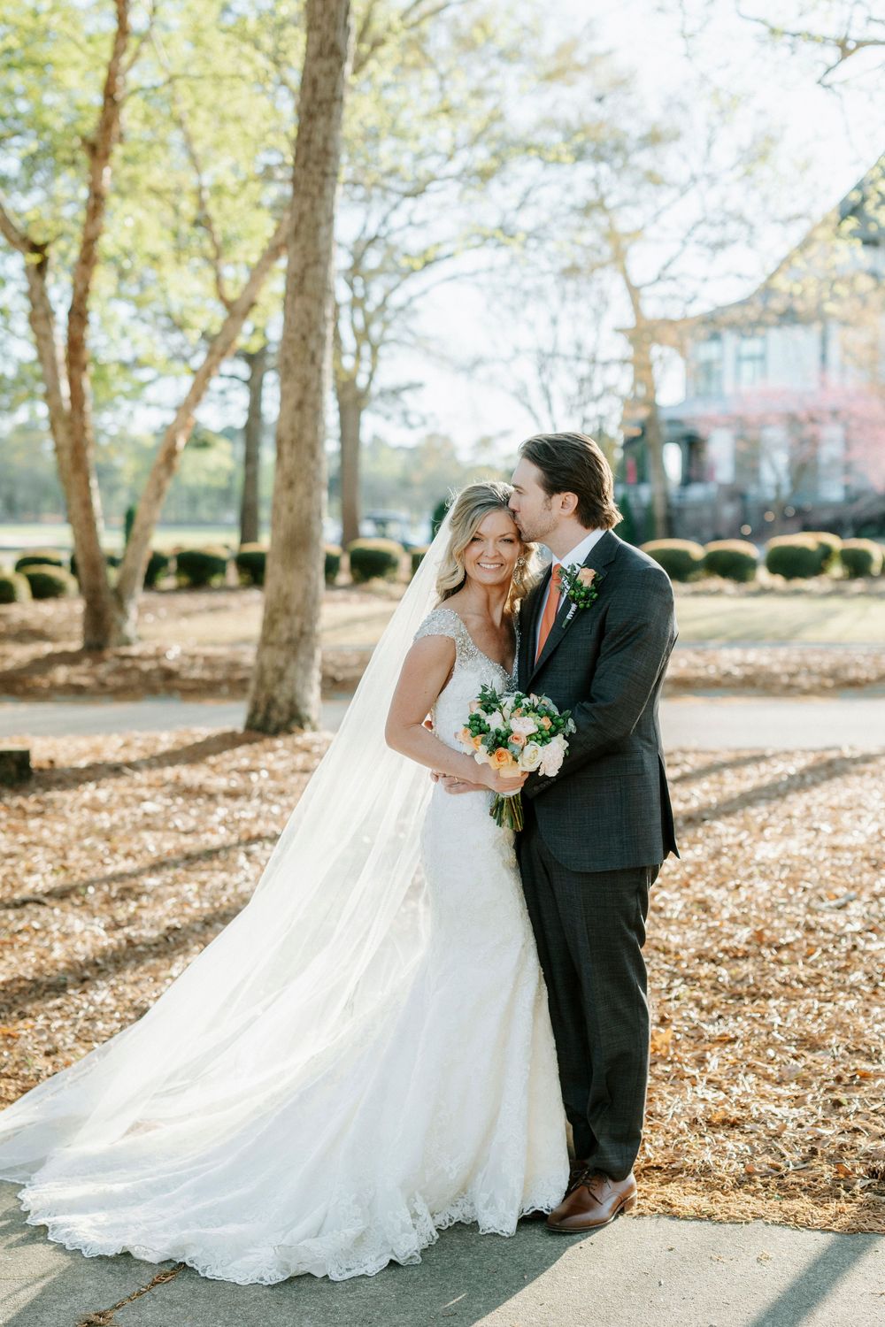 Romantic wedding couple sharing a tender moment on a sunlit path surrounded by autumn leaves and trees.