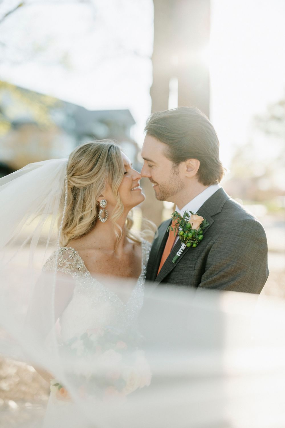 Newlyweds share a tender moment outdoors with sunlight filtering through a delicate white veil.