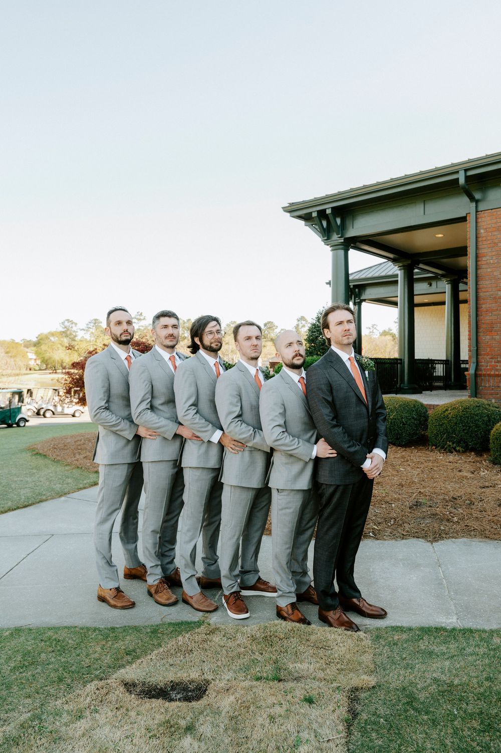 Groomsmen lined up outside venue wearing matching light gray suits with brown shoes while best man wears black suit.