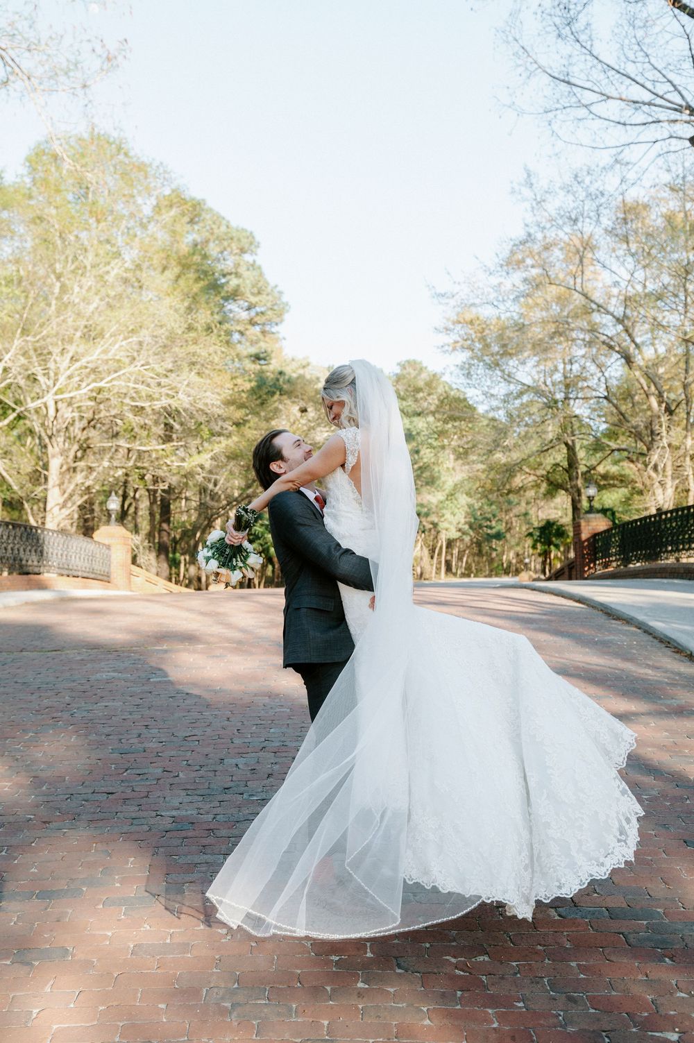 Wedding couple dances and twirls on a brick patio surrounded by trees at dusk.