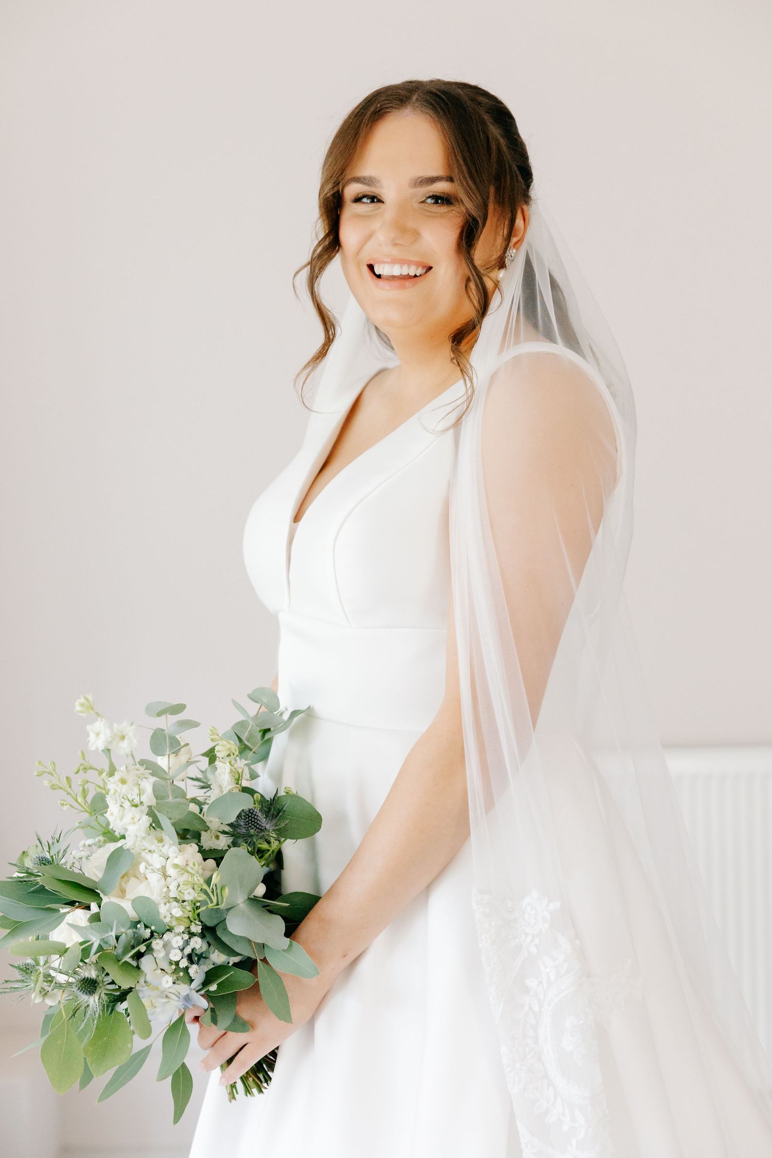 A bride in a white wedding dress poses with a lush green and white eucalyptus bouquet against a light backdrop at Wickham Estate