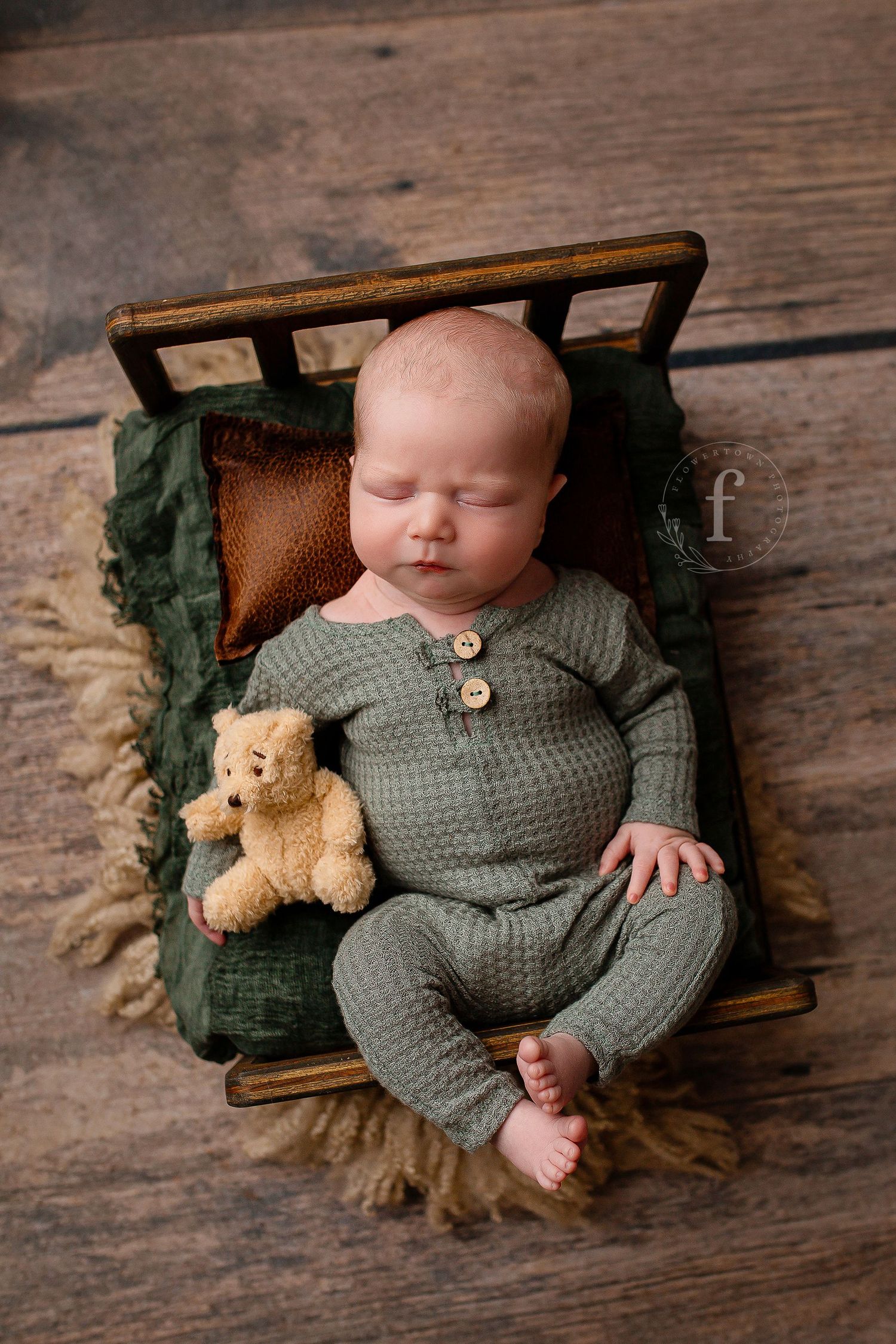Older newborn sleeping in green outfit with teddy bear on small bed on hardwood floor in Flowertown Photography Studio in Summerville, SC