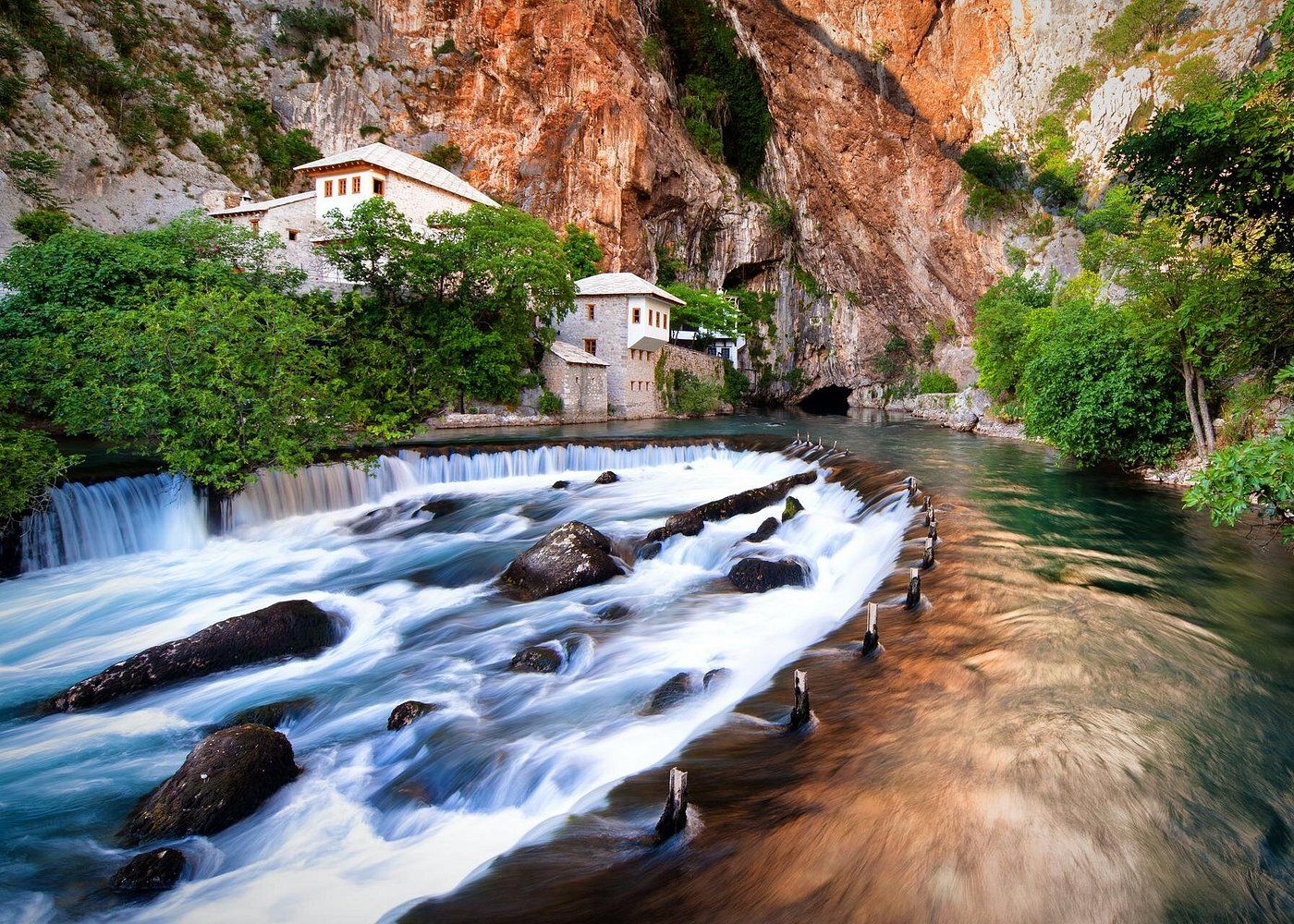 Cascading waterfall flows through rocky canyon with white building nestled in cliff face above rushing mountain stream.