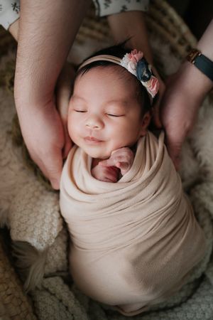 Newborn baby swaddled in peach wrap with floral headband, sleeping peacefully while parents cradle her head.新生兒裹在蜜桃色襁褓裡，戴著花朵髮帶，父母輕輕托著她的頭，她睡得