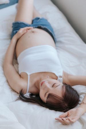 Expectant mother in white crop top and denim relaxes peacefully on white bedding穿著白色露臍上衣和牛仔褲的準媽媽平靜地躺在白色床單上。