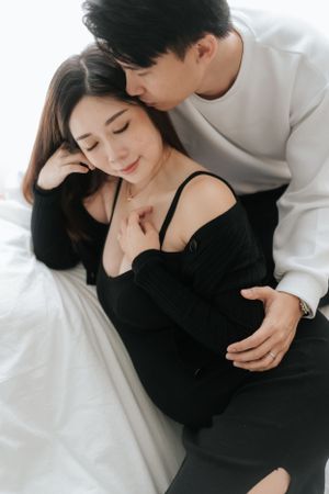Intimate couple embraces on white bedding as they share a tender moment in soft lighting.一對情侶在白色床單上親密相擁，在柔和的燈光下共享溫馨時刻。