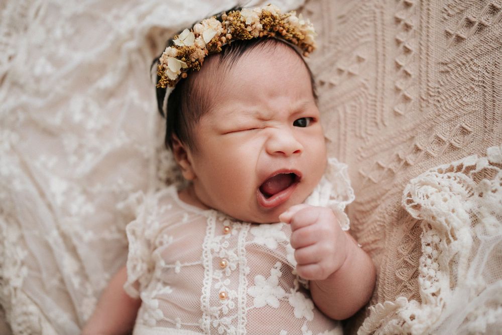 Baby wearing lace dress and floral crown yawning on textured blanket.穿著蕾絲洋裝、戴著花冠的嬰兒在有紋理的毯子上打哈欠。