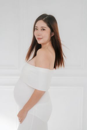 Elegant maternity portrait in white off-shoulder dress against light background.優雅的孕婦照，穿著白色露肩連身裙，背景為淺色。