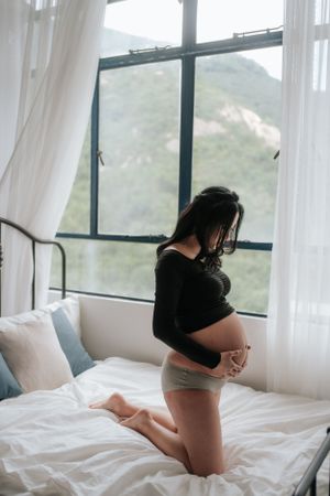Silhouette of pregnant woman by window with mountain view, kneeling on white bed.窗外山景中，一位孕婦跪在白色床上，身影若隱若現。