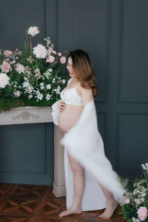 Elegant maternity photoshoot with white dress against dark paneled wall and pink flowers.優雅的孕婦寫真，身穿白色連身裙，背景是深色鑲板牆和粉紅色鮮花。
