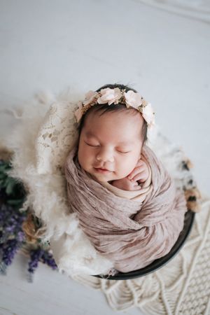 Sleeping newborn with floral crown wrapped in soft fabric against white backdrop.睡著的新生兒頭戴花冠，全身裹在柔軟的布料裡，背景是純白色。