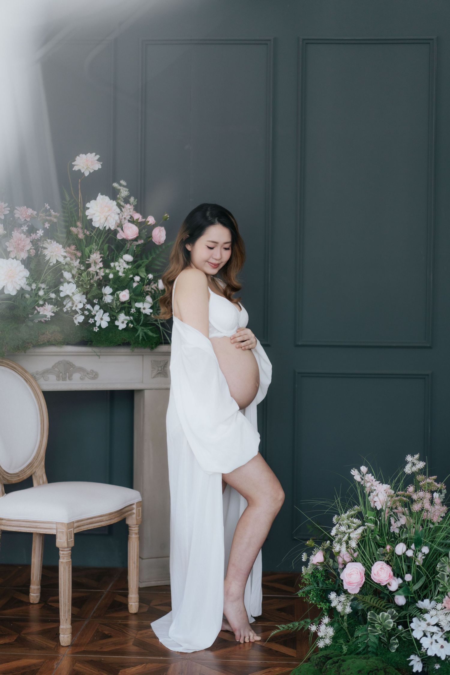 Artistic portrait with flowing white fabric against dark wall with pink floral arrangements.藝術肖像畫，白色飄逸的布料襯托著深色牆面和粉紅色花卉裝飾。