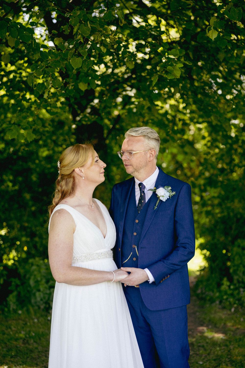 Bride and groom gaze lovingly at each other holding hands beneath lush green trees on wedding day.
