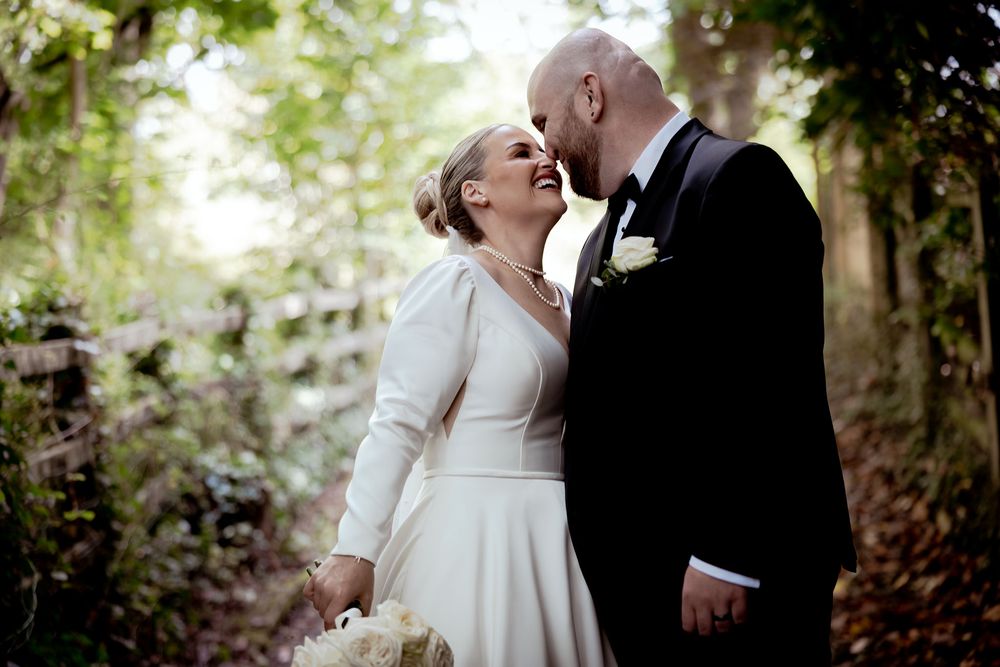 Bride and groom share a romantic moment in a lush forest setting, with the groom leaning in for a kiss. village hotel Cardiff
