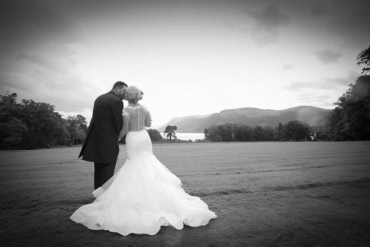 Black and white wedding photo of bride and groom embracing in open field with mountain backdrop.
