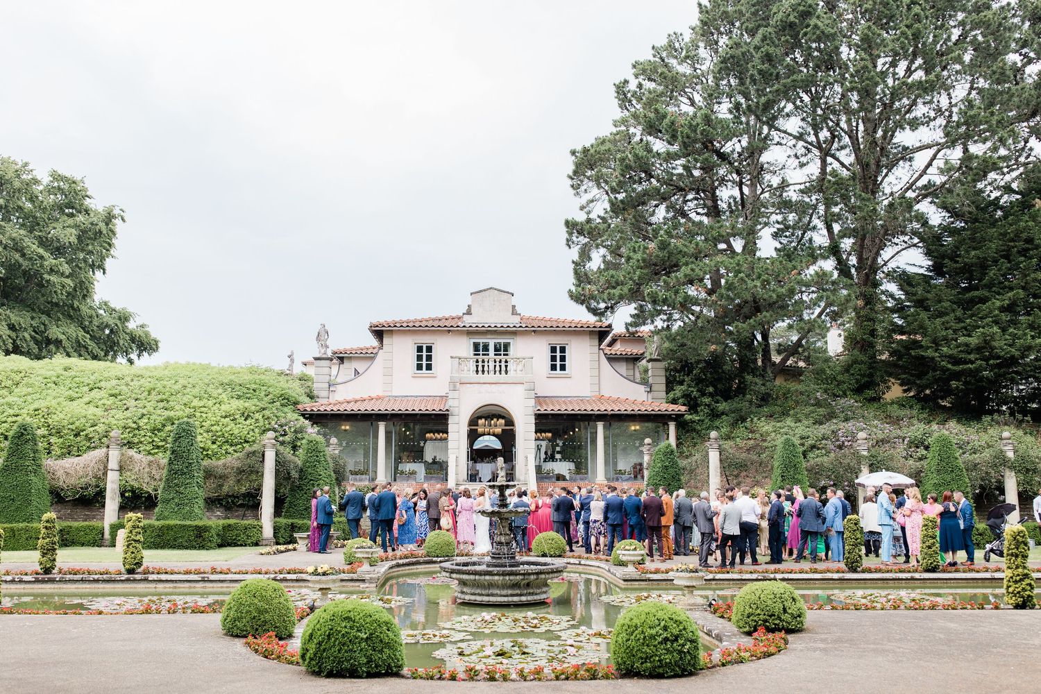 Wedding guests gather outside the Italian Villa with stone walls and manicured gardens on a sunny afternoon by Maddie Waters