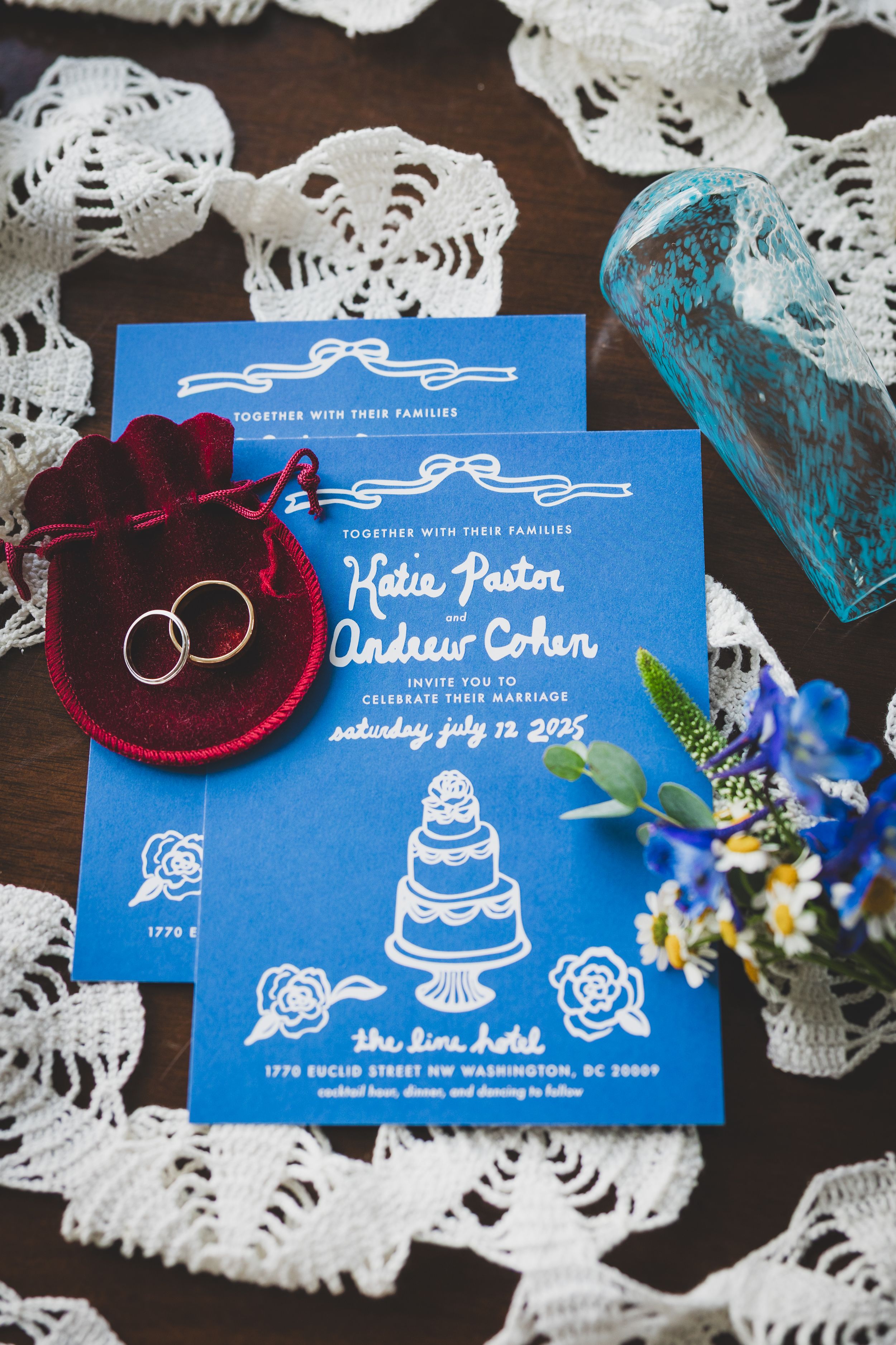 Blue wedding invitation cards with white lace doilies and decorative wedding rings displayed on dark wooden surface.