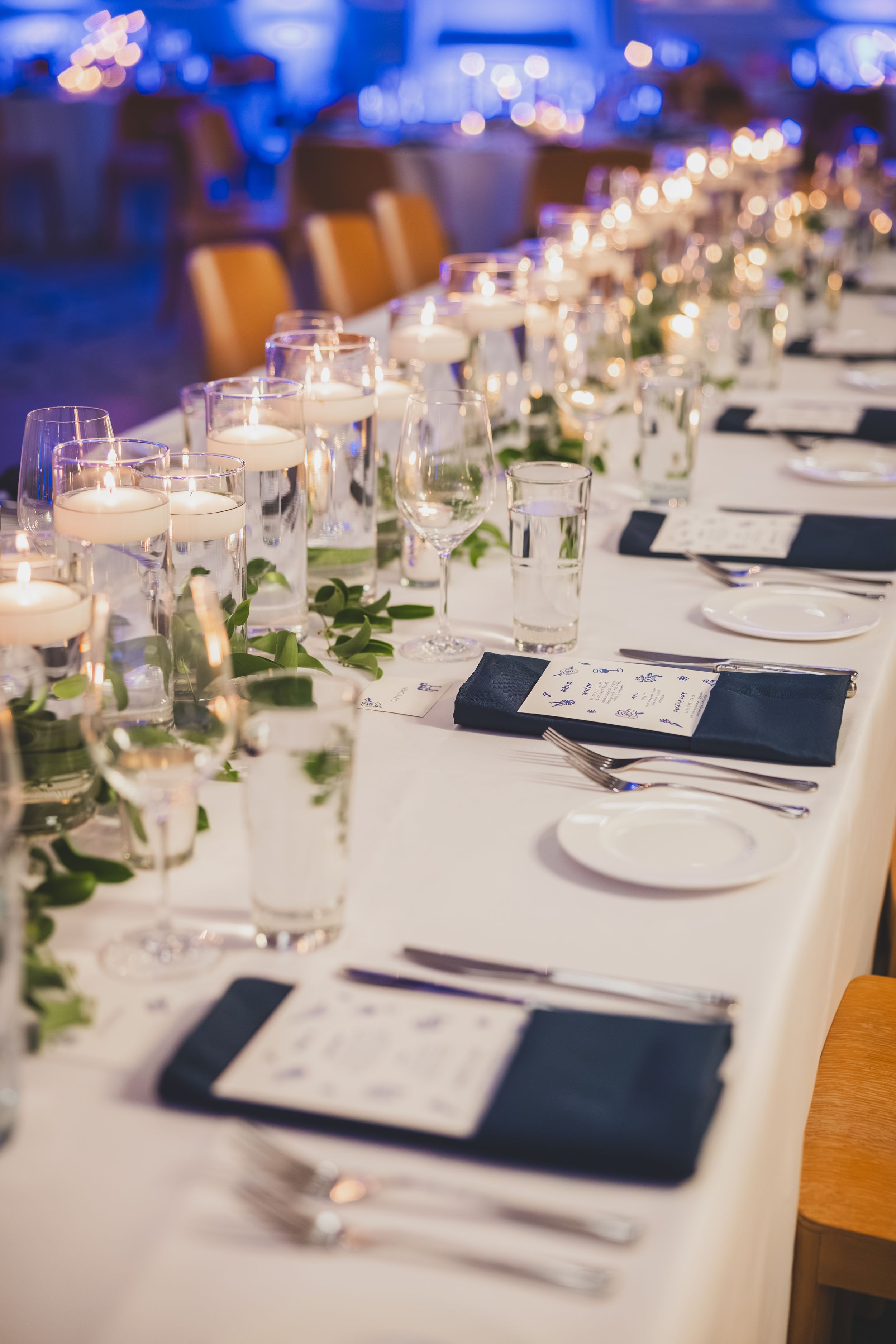 Elegant wedding reception table settings with white linens, navy blue napkins, and glassware in soft blue lighting.
