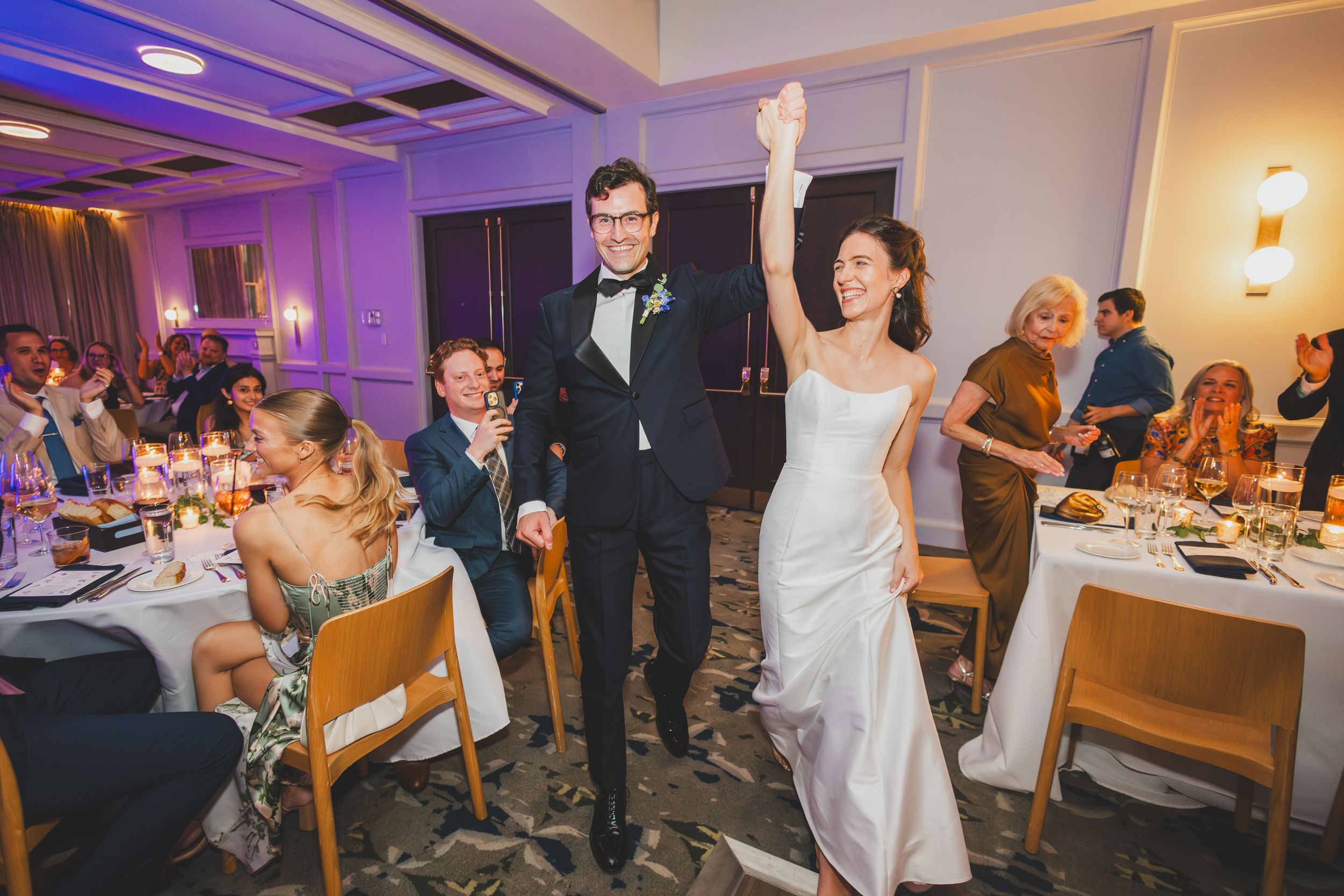 Wedding celebration at reception venue with couple making grand entrance into purple-lit dining room with cheering guests.
