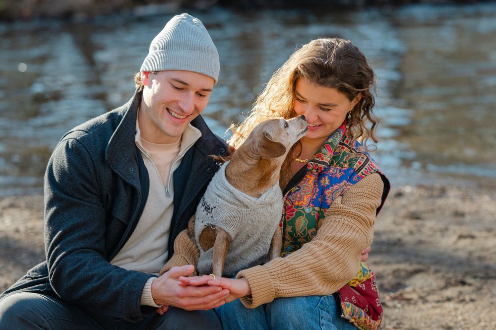 Two people sitting by the water looking at a phone together while wearing casual winter clothing on a sunny day.