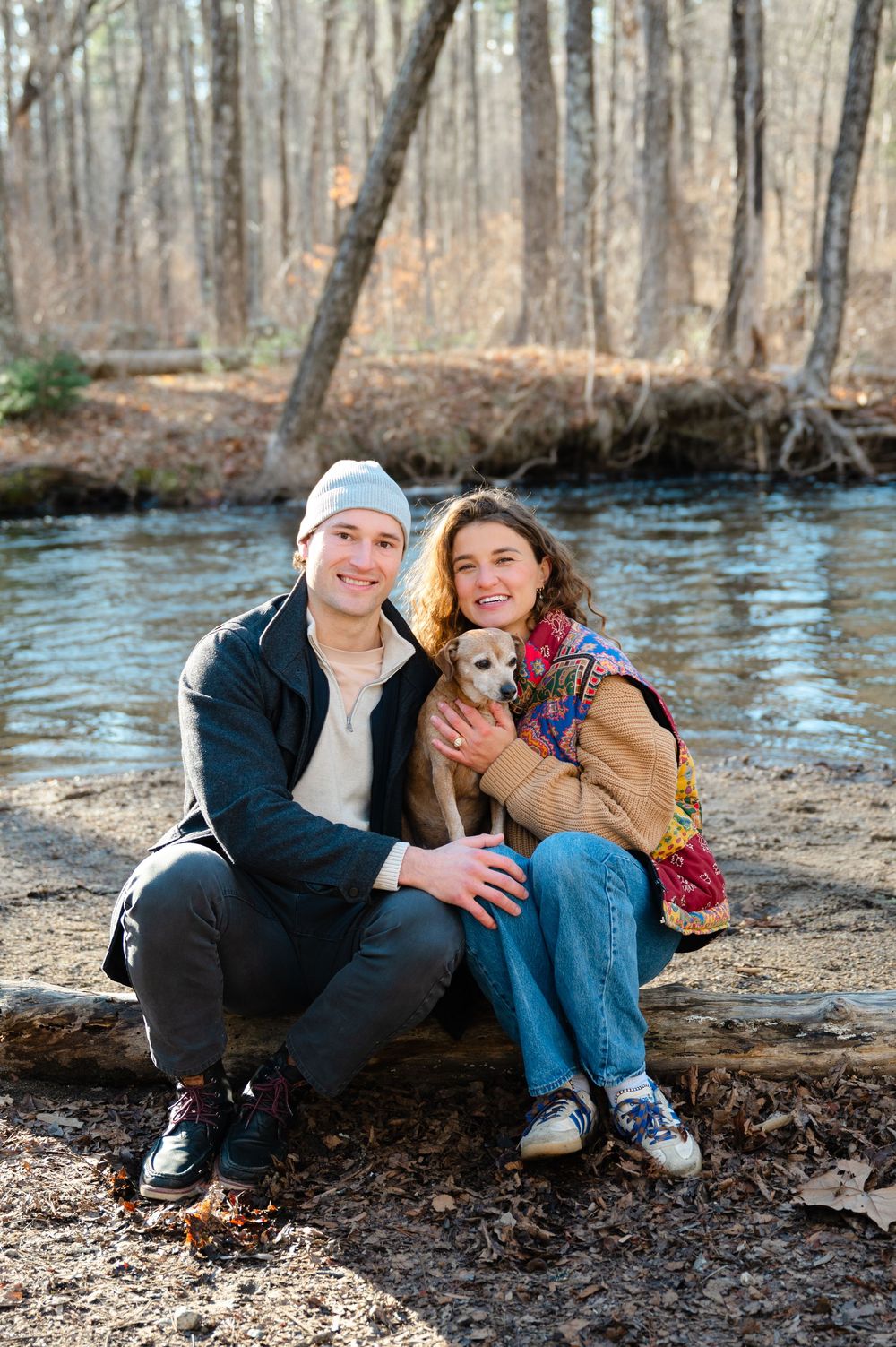 A couple sits with their small dog by a stream in the woods on a sunny winter afternoon.