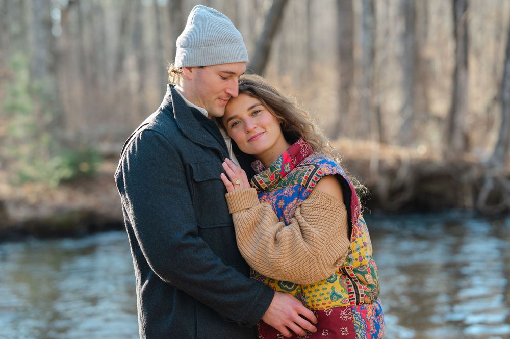 A couple embraces by a river in winter, wearing warm clothing including a beanie and colorful scarf.