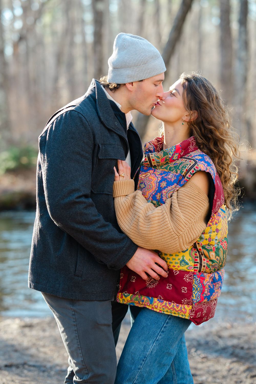 A romantic couple sharing a kiss outdoors by a stream, wearing winter clothing and a colorful patterned vest.
