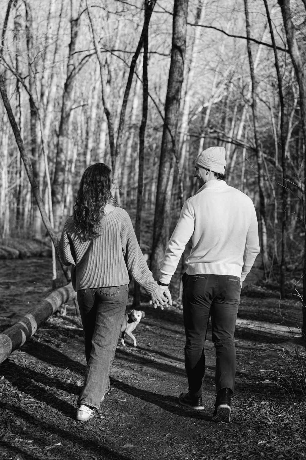 A couple walks hand in hand with their dog along a wooded trail on a winter day, captured in black and white.