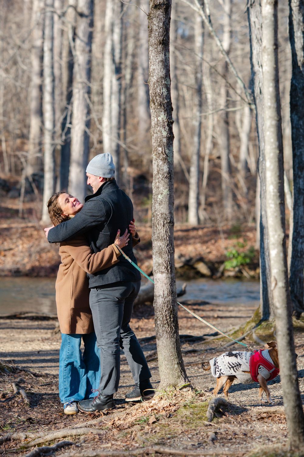 A couple shares a romantic moment in a wooded area by a stream while their dog watches nearby.