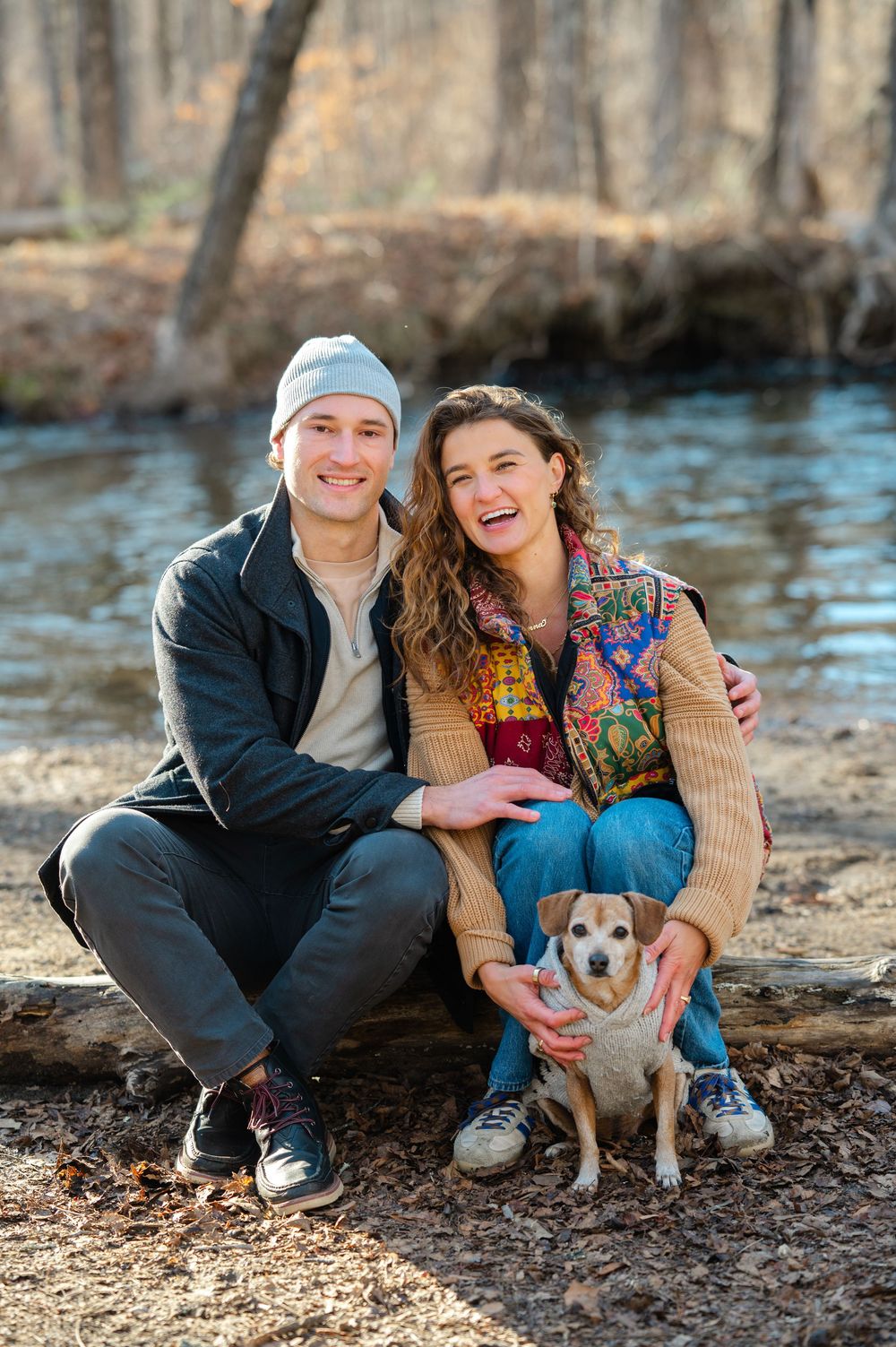A couple sits with their small dog by a river on a sunny day, surrounded by bare trees in Ward Pound Ridge Reservation after their proposal.
