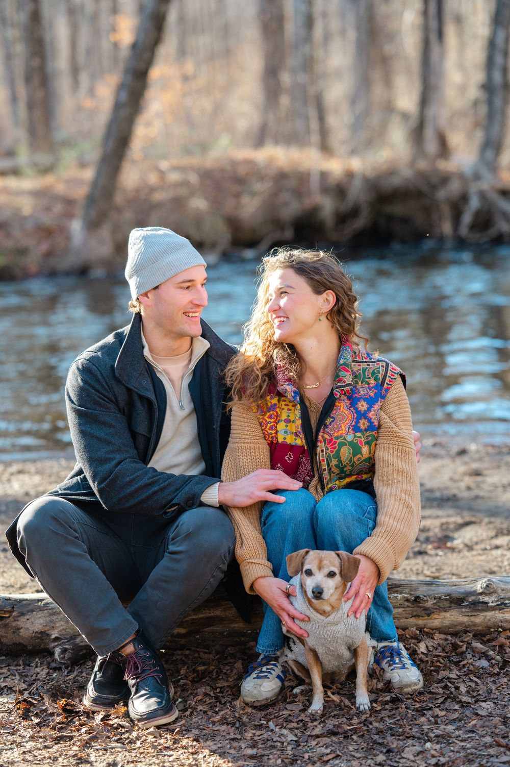 A couple sits on fallen leaves by a stream with their small dog while sharing an intimate moment in nature.