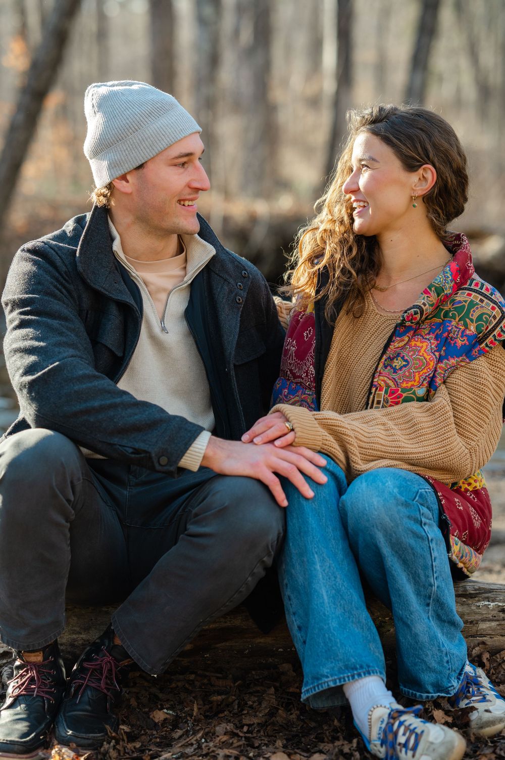 A couple sits together on a fallen log in the woods, wearing casual winter clothing and sharing a tender moment.