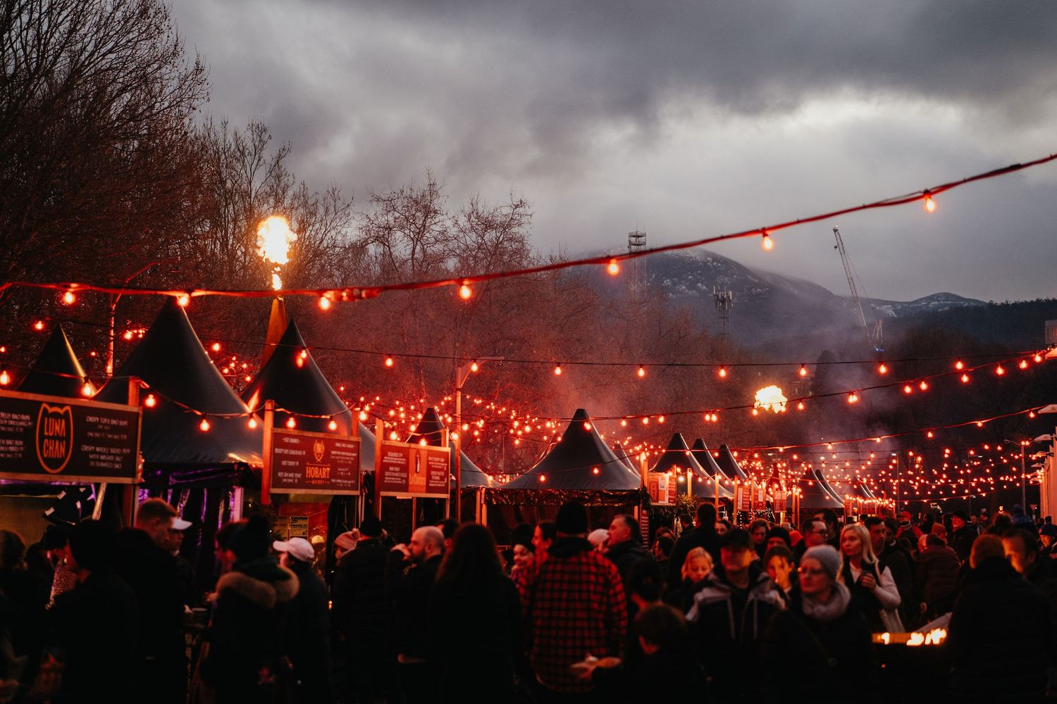 A large outdoor crowd gathers under red string lights at dusk during a festive evening event.