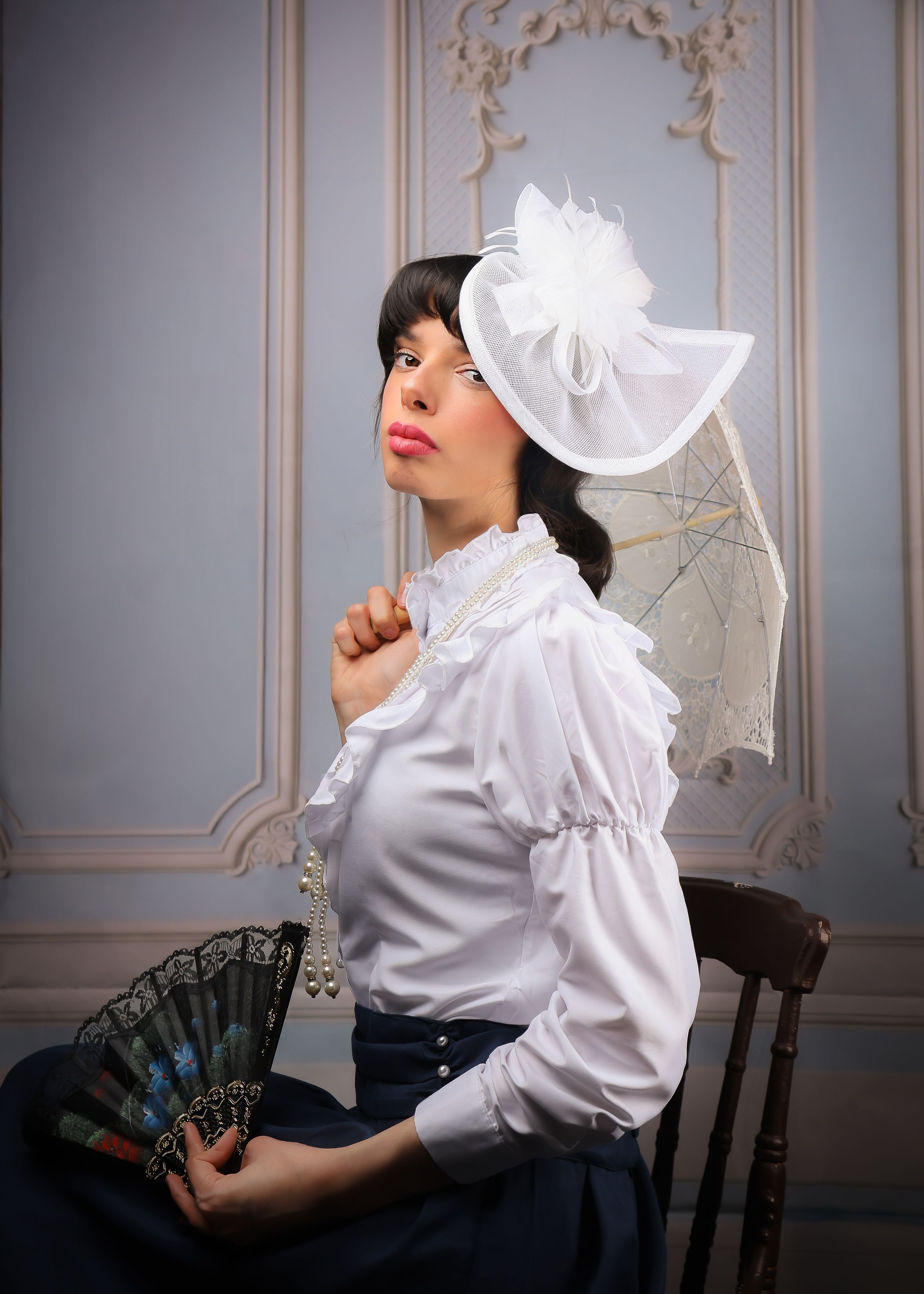 Photo of a girl in a Victorian, edwardian dress - holding a parasol, and a regency hat - looking at the camera from left angle