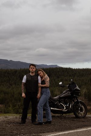 Tattooed couple standing together on a gravel road with a black motorcycle and forested mountains behind taken by Yelm WA photographer