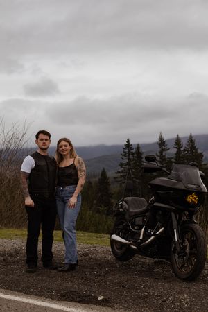 Couple posing beside a black Harley-Davidson motorcycle on a scenic overlook with mountains and cloudy skies in Yelm Washington