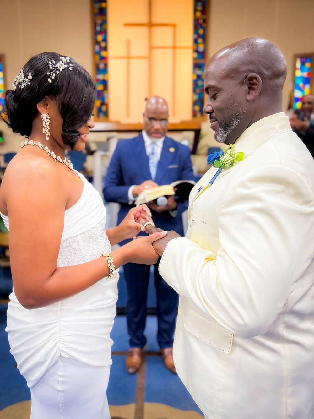 Bride placing wedding ring on groom's finger during church ceremony, officiant holding Bible in background.