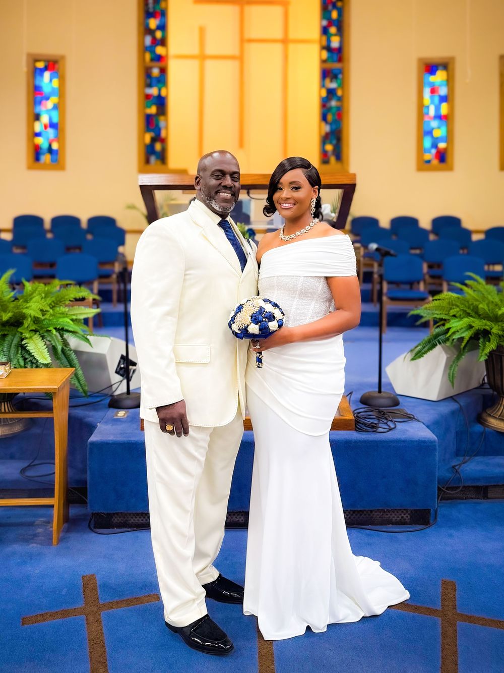 Bride and groom posing in white wedding attire inside a church with blue chairs and stained glass windows.