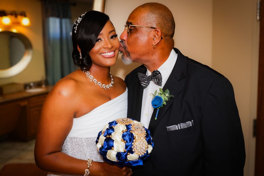 A groom kisses the cheek of his smiling bride holding a blue and white bouquet on their wedding day.
