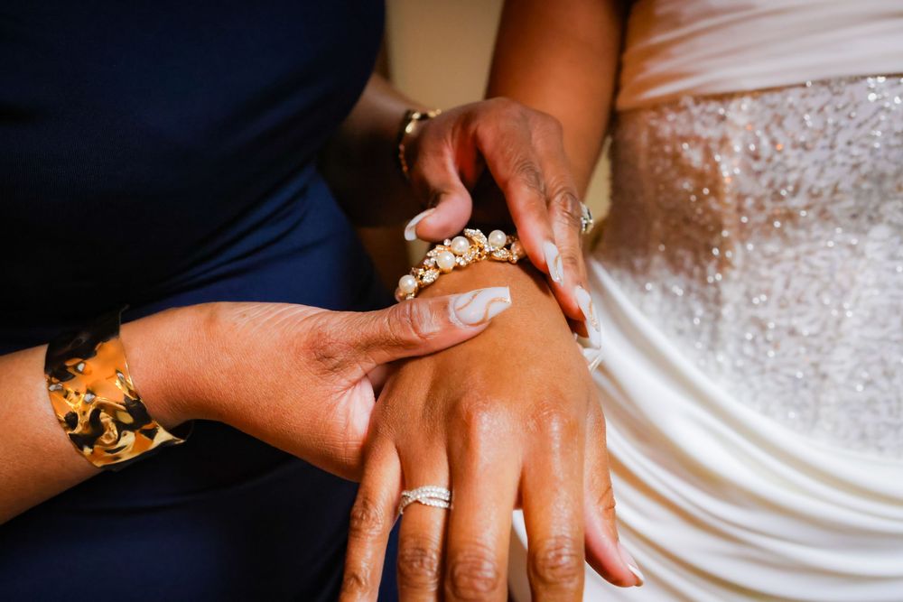 Two women in elegant attire hold hands, showcasing gold bracelets and rings at a wedding ceremony.