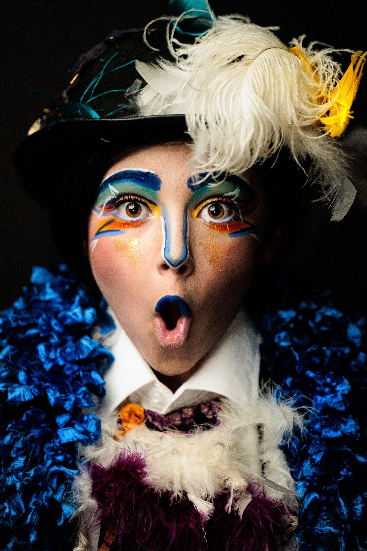 Theater performer with colorful bird-inspired face paint, feathered hat, and blue ruffled costume.