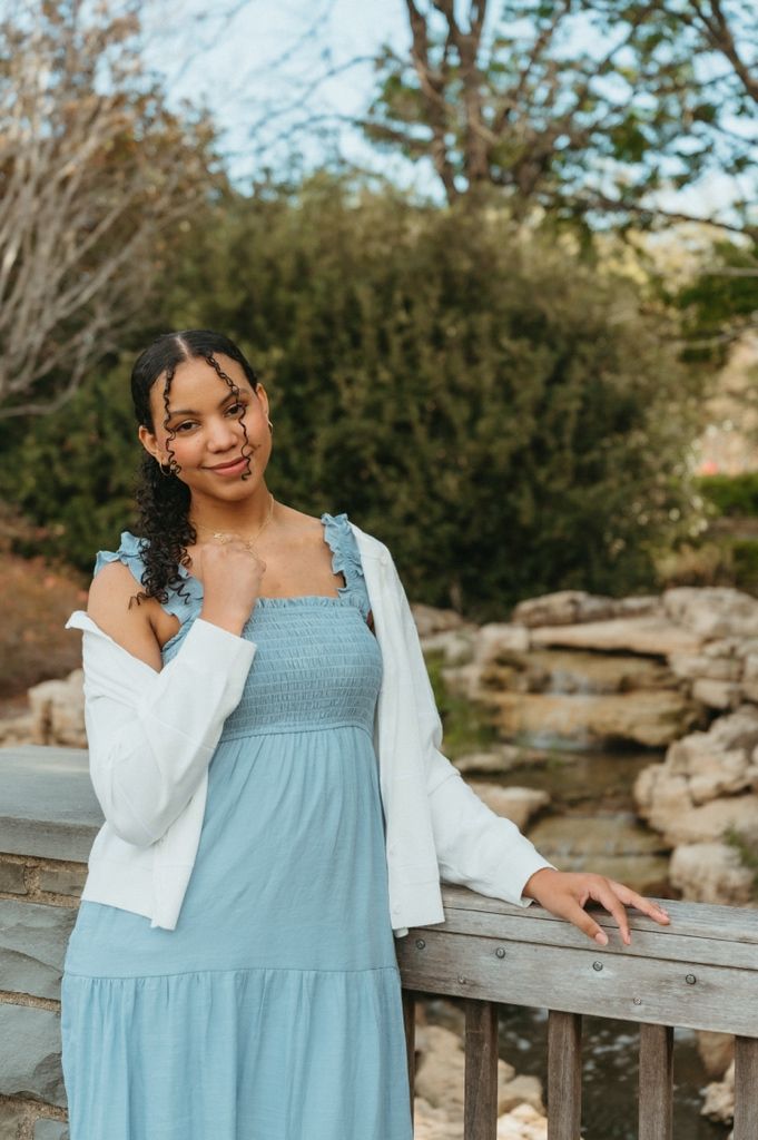 Young woman in light blue smocked dress and white jacket poses by wooden railing in outdoor garden setting.