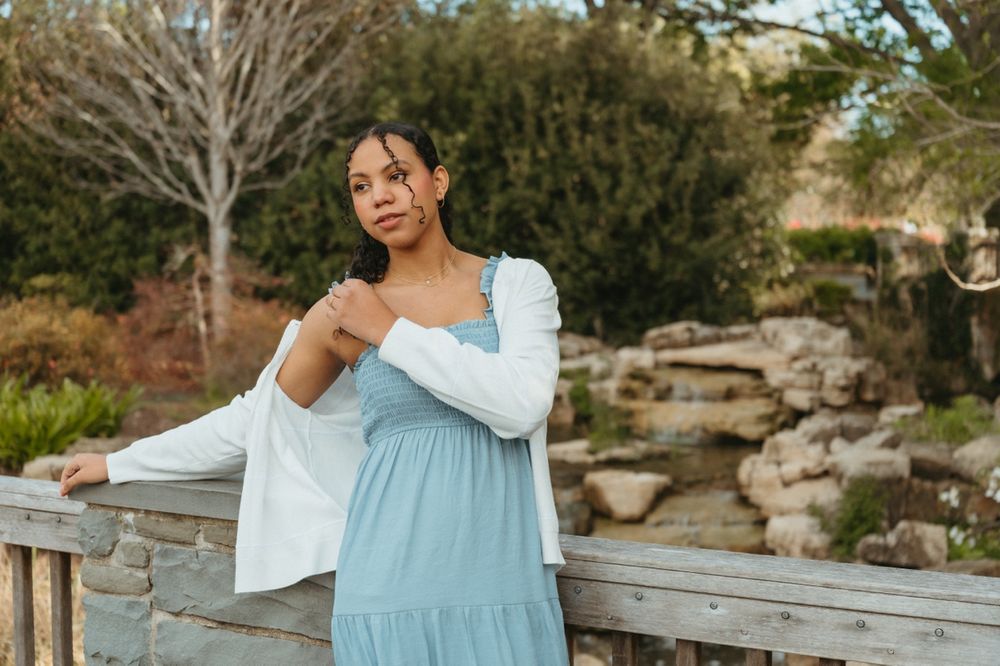 Woman in light blue maxi dress and white cardigan posing outdoors near wooden fence and stone wall.