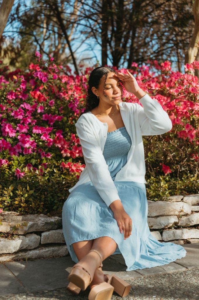 Woman in light blue dress and white cardigan sitting on stone wall surrounded by vibrant pink flowers.