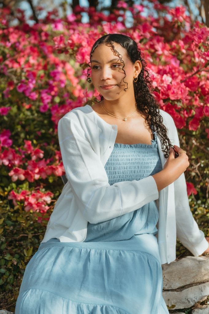 Young woman in light blue dress and white cardigan sitting near vibrant pink flowers.