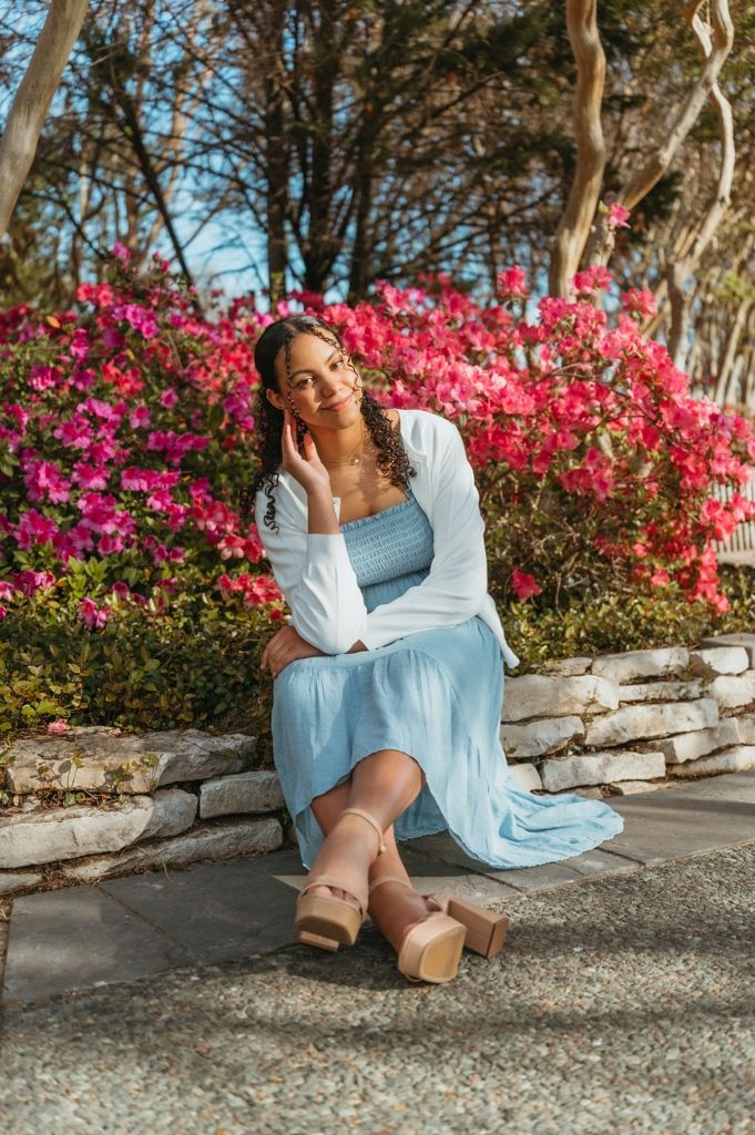 Woman in blue dress and white cardigan seated on stone wall with pink blooms behind her.