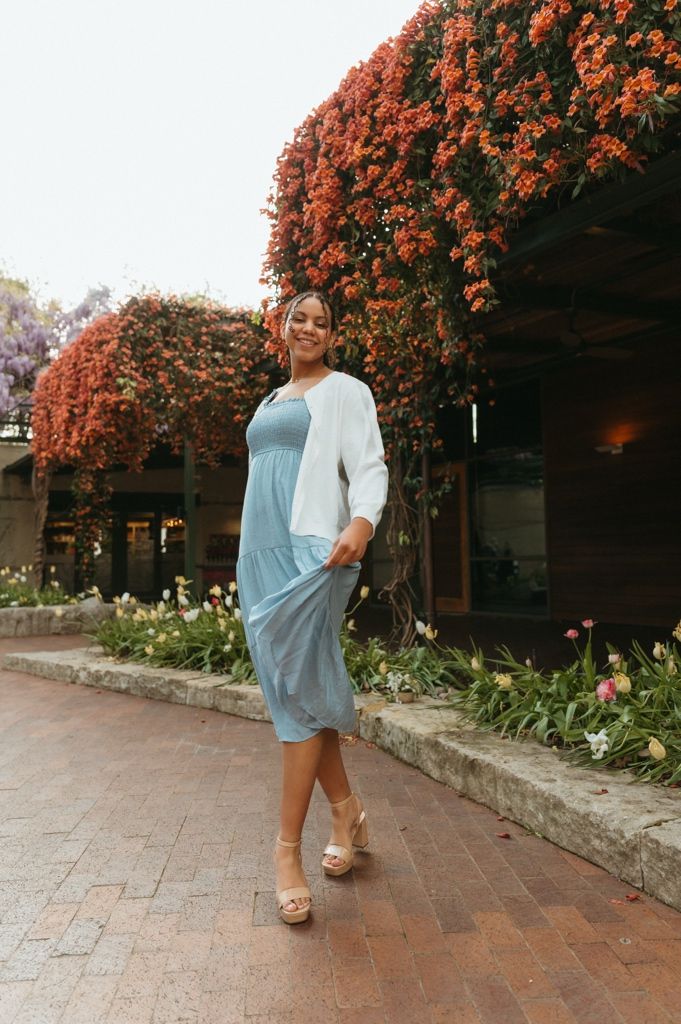 Woman in blue midi dress and white cardigan posing on brick path with vibrant red flowering wall behind her.