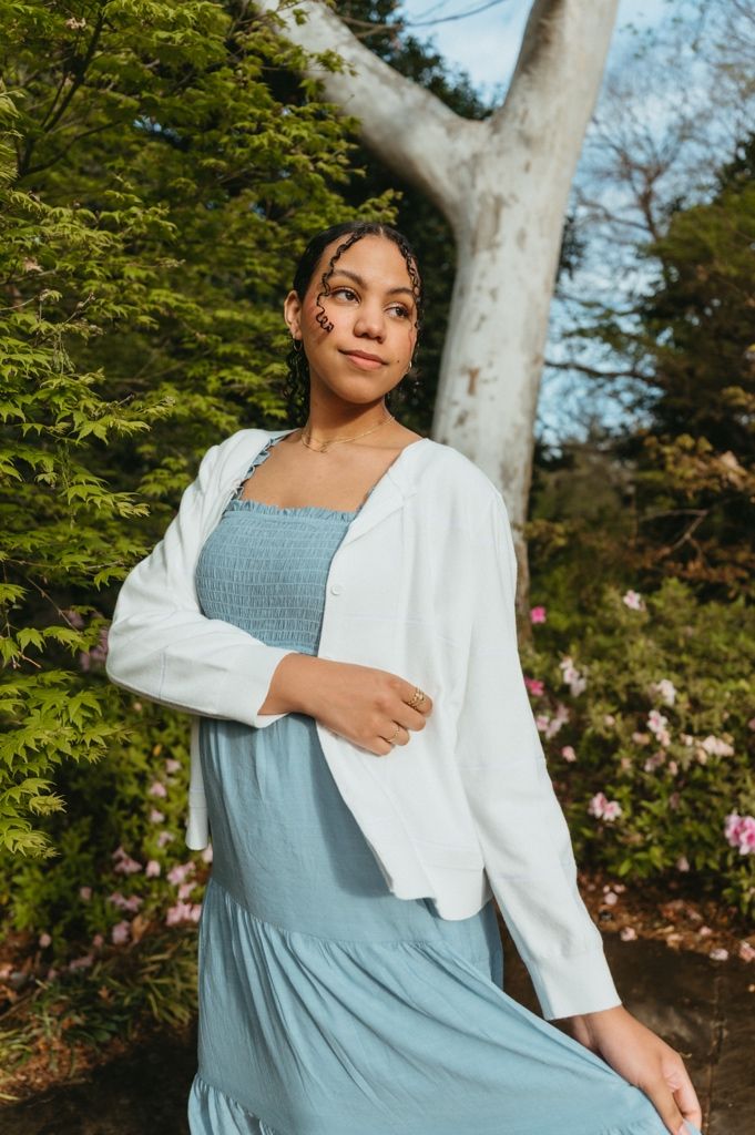 Young woman in light blue dress and white cardigan poses outdoors near trees with pink flowers in bloom.