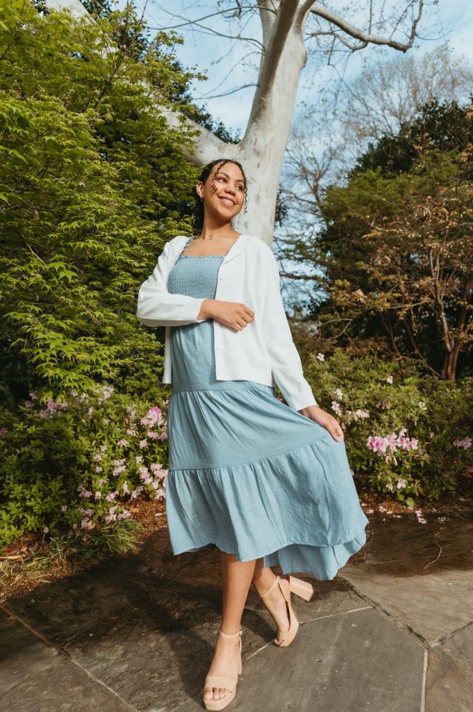Woman in light blue tiered midi dress and white cardigan poses outdoors near flowering shrubs and trees.